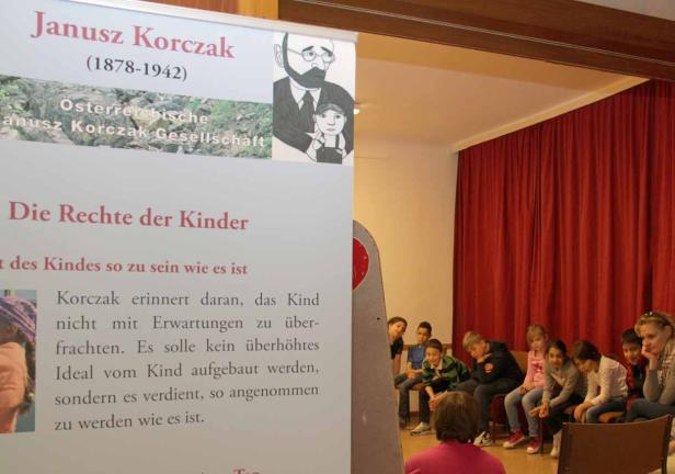 Eine Gruppe Kinder sitzt vor einem Plakat über Janusz Korczak und die Rechte der Kinder.