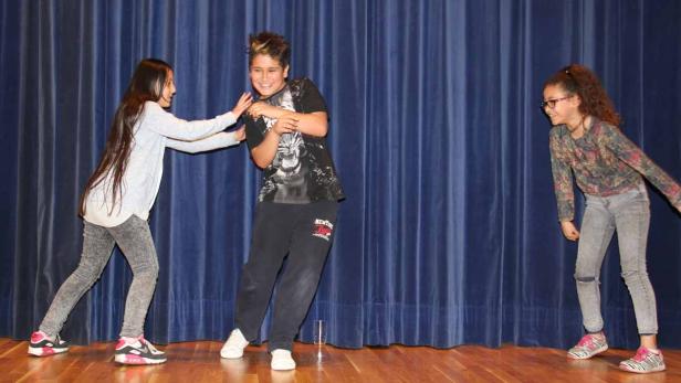 Drei Kinder stehen auf einer Bühne vor einem blauen Vorhang und scheinen ein Theaterstück aufzuführen.