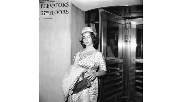 Elizabeth Taylor mit Hut und Kleid vor einem Aufzug.