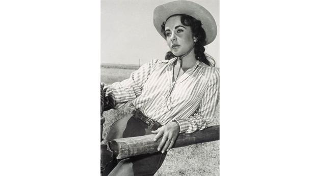Elizabeth Taylor in Cowboy-Kleidung, lässig an einem Zaun lehnend.