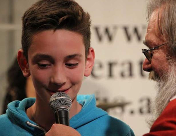 Ein lächelnder Junge spricht in ein Mikrofon, während ein älterer Mann mit Bart daneben steht.