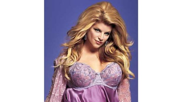 Kirstie Alley trägt ein lila Negligé und posiert vor einem blauen Hintergrund.