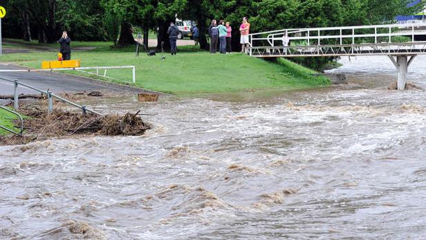 Hochwasser überflutet eine Straße und einen Fluss, während Menschen auf einer Brücke stehen.
