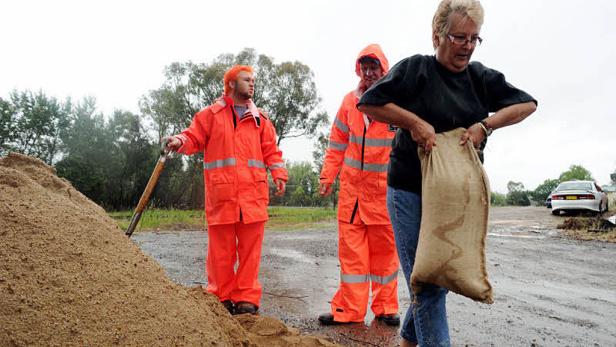 Drei Personen füllen Sandsäcke neben einem Sandhaufen, möglicherweise als Schutz vor Überschwemmungen.