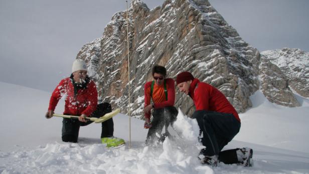 Drei Personen schaufeln Schnee vor einer schneebedeckten Bergkulisse.