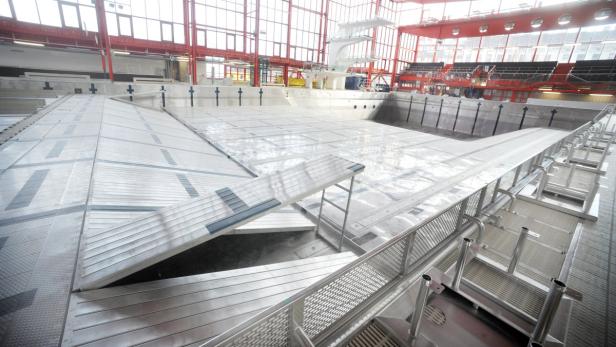 Ein leeres Schwimmbecken mit einer temporären, silbernen Abdeckung und einem Sprungturm im Hintergrund.