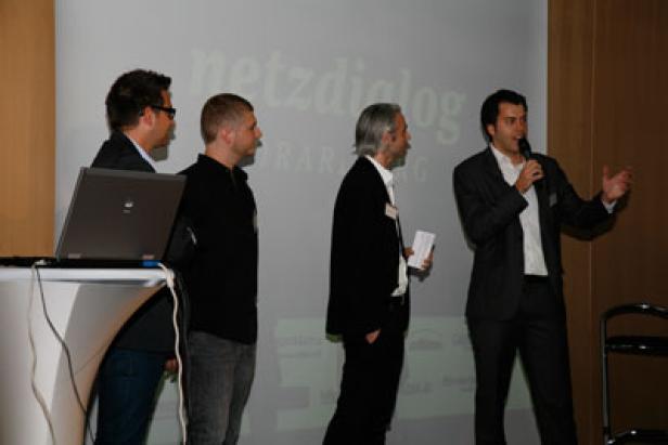Ein Mann hält ein Mikrofon und spricht vor einer Gruppe von Personen bei einer „netzdialog“-Veranstaltung.