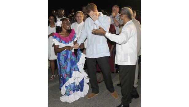 Prinz Harry tanzt mit einer Frau in traditioneller Kleidung, während ein Mann ihm etwas erklärt.