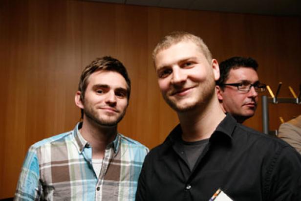 Zwei Männer posieren für ein Foto, ein dritter Mann ist im Hintergrund zu sehen.