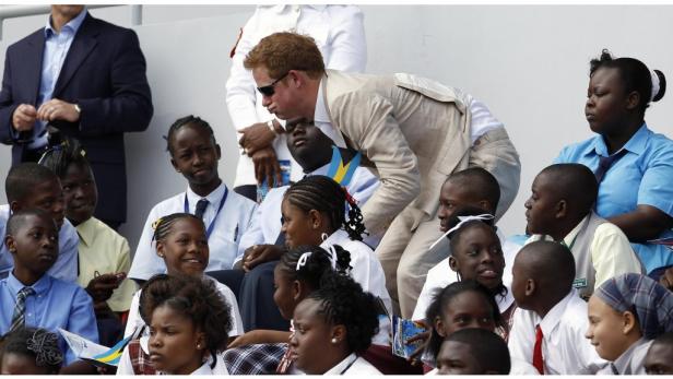 Prinz Harry beugt sich zu einer Gruppe von Schulkindern auf einer Tribüne.