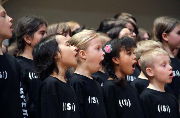 Eine Gruppe Kinder singt in einem Chor, gekleidet in schwarzen T-Shirts mit einem weißen Symbol.
