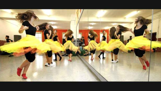 Eine Gruppe junger Tänzerinnen in gelben Tutus übt in einem Tanzstudio vor einem Spiegel.