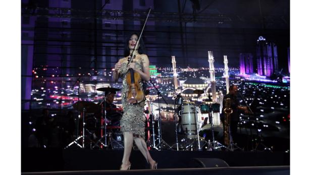 Eine Geigerin tritt mit ihrer Band vor einer beleuchteten Stadtkulisse auf.