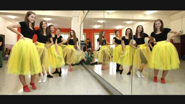 Eine Gruppe junger Frauen in schwarzen Oberteilen und gelben Tutus posiert in einem Tanzstudio.