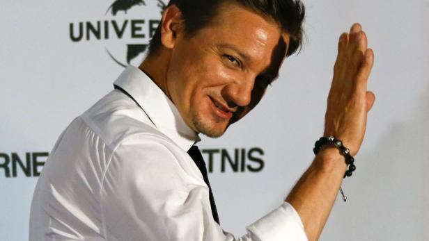 Jeremy Renner lächelt und winkt in einem weißen Hemd und mit Armband.