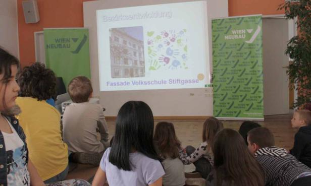 Kinder sitzen vor einer Präsentation über die Fassade der Volksschule Stiftgasse.