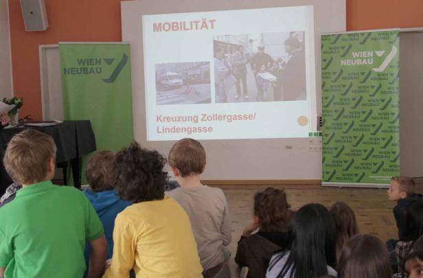 Kinder sitzen vor einer Leinwand mit der Aufschrift „Mobilität“ und dem Text „Kreuzung Zollergasse/Lindengasse“.