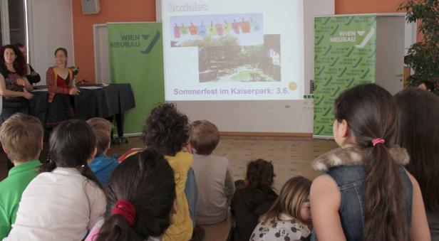 Kinder sitzen vor einer Leinwand, auf der ein Sommerfest im Kaiserpark angekündigt wird.