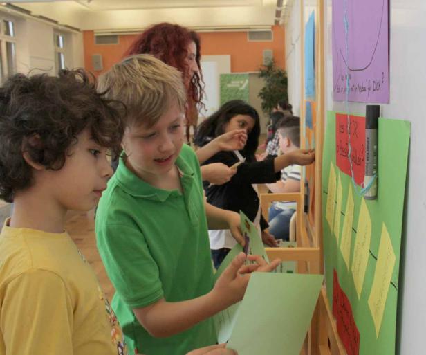 Zwei Jungen betrachten Zettel an einer Wand, an der weitere Personen arbeiten.