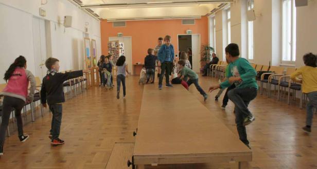 Eine Gruppe Kinder spielt in einem hellen Raum auf und neben einer langen Tischtennisplatte.