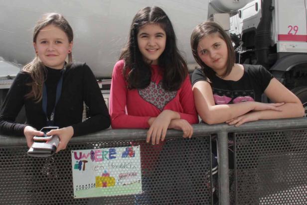 Drei Mädchen stehen lächelnd an einer Absperrung vor einem LKW.