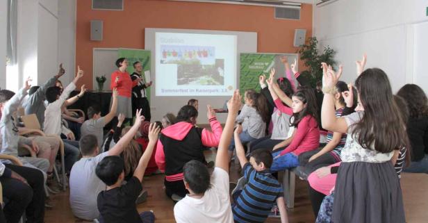 Eine Gruppe von Schülern sitzt in einem Klassenzimmer und hebt die Hände, während zwei Lehrer vorne stehen.