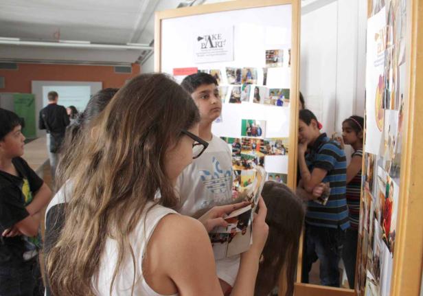 Eine Gruppe von Kindern betrachtet eine Pinnwand mit Fotos und dem Slogan „Take Part!“.