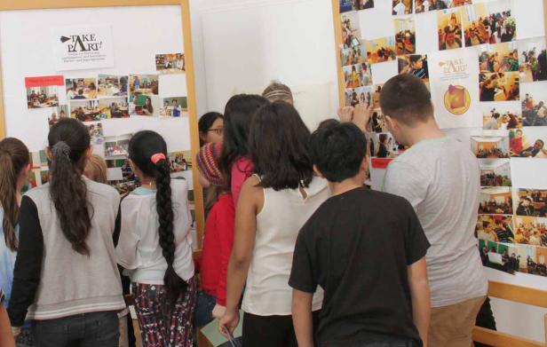Eine Gruppe von Jugendlichen betrachtet eine Pinnwand mit Fotos und dem Logo „Take Part!“.