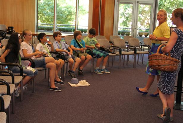 Eine Gruppe von Kindern sitzt in einem Raum, während zwei Frauen stehen und sich unterhalten.