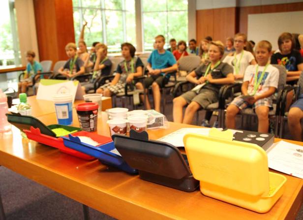 Kinder sitzen in einem Raum vor Tischen mit Bastelmaterialien.