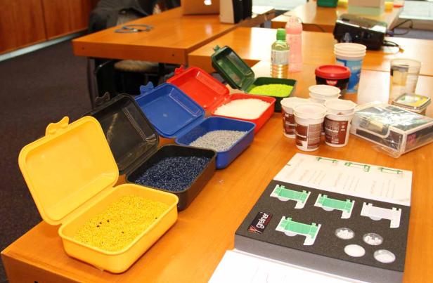 Auf einem Tisch stehen Behälter mit farbigen Kunststoffgranulaten und Verpackungsmaterialien.