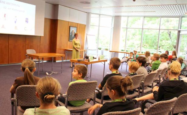 Eine Frau hält einen Vortrag vor einer Gruppe von Kindern in einem hellen Raum.