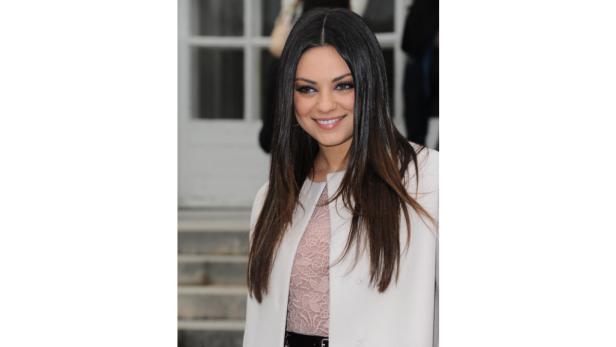 Mila Kunis lächelt in einem weißen Mantel und einem rosa Spitzenoberteil.