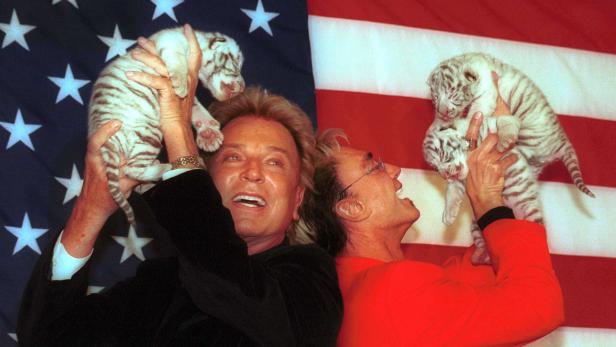 Siegfried und Roy halten jeweils ein junges weißes Tigerbaby vor einer US-Flagge hoch.