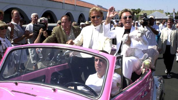 Siegfried und Roy fahren in einem pinkfarbenen Auto, Roy hält ein junges weißes Tier im Arm.