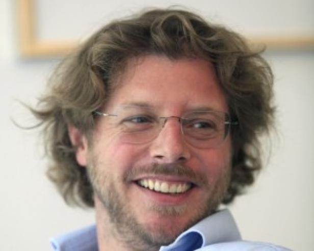 Ein Mann mit lockigem Haar und Brille lächelt in die Kamera.