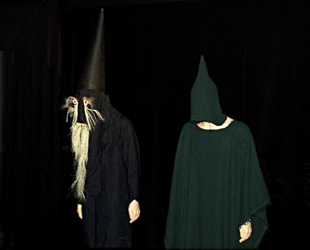 Zwei Personen in ungewöhnlichen Kostümen stehen vor einem schwarzen Hintergrund.