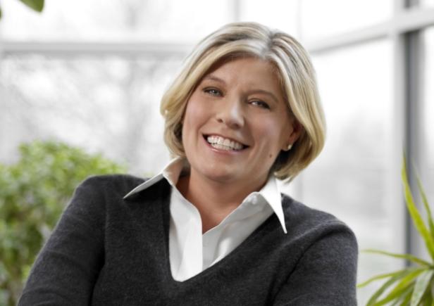 Eine lächelnde Frau mit blonden Haaren und einem grauen Pullover vor einem Fenster.