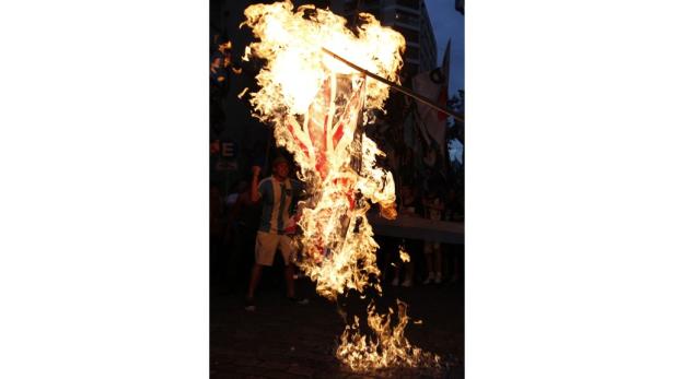 Eine britische Flagge steht in Flammen, während eine Menschenmenge im Hintergrund steht.