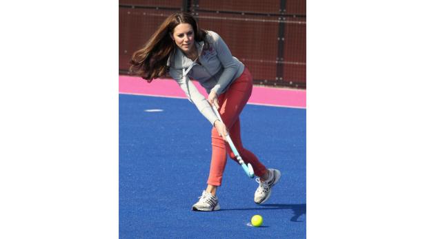 Kate Middleton spielt Hockey auf einem blauen Feld.