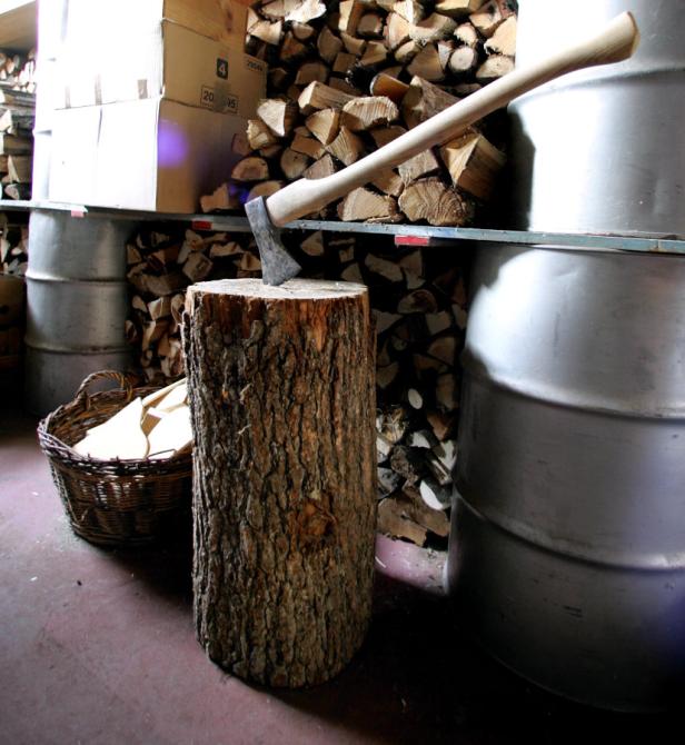 Ein Holzspalter mit Axt steht vor einem Regal mit Brennholz und Fässern.
