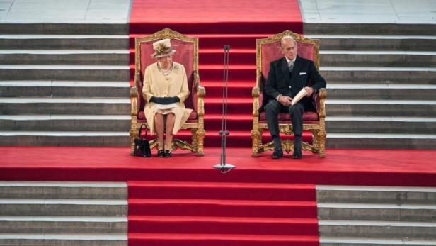 Königin Elisabeth II. und Prinz Philip sitzen auf Thronen vor einer Treppe mit rotem Teppich.