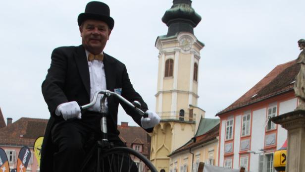 Ein Mann in Frack und Zylinder fährt auf einem Hochrad durch eine Stadt.