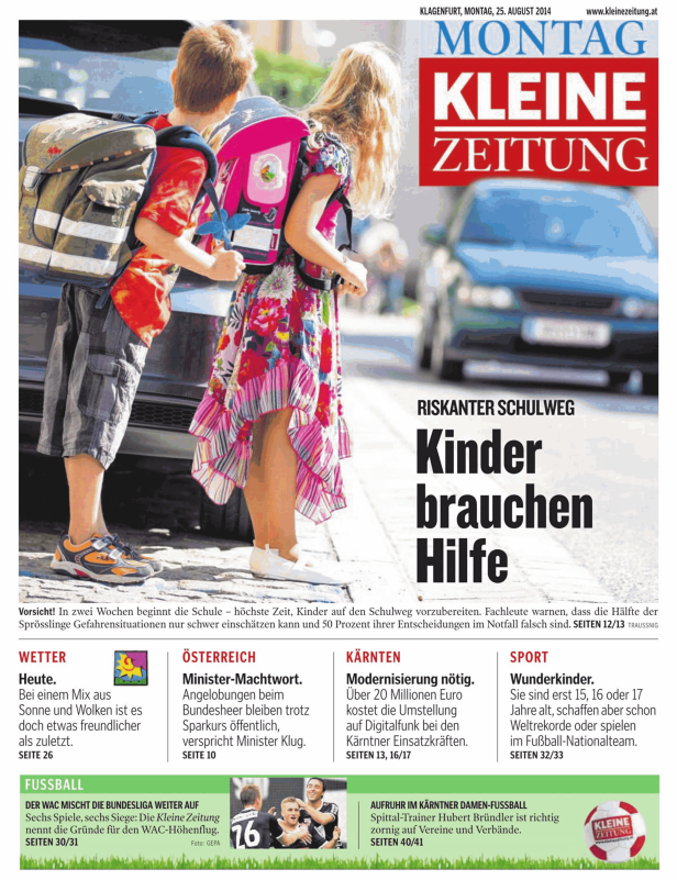 Zwei Kinder mit Schulranzen stehen an einer Straße.