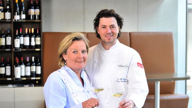 Eine Frau und ein Koch halten Weingläser vor einem Regal mit Weinflaschen.