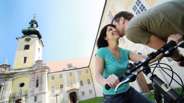 Ein Paar auf Fahrrädern vor dem Stift Herzogenburg, wobei der Mann die Frau auf die Wange küsst.