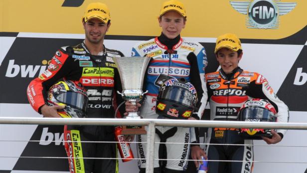 Drei Rennfahrer stehen mit Helmen und einem Pokal auf dem Siegerpodest.