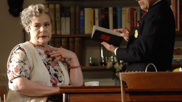 Eine ältere Dame sitzt an einem Tisch, während ein Mann ihr ein Buch zeigt.