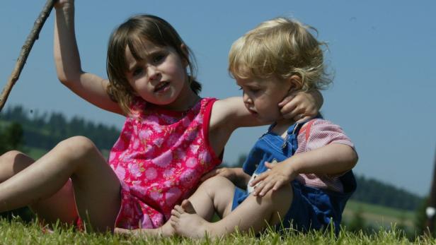 Ein Mädchen mit einem Stock sitzt neben einem kleineren Kind im Gras.