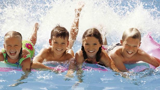 Vier lachende Kinder planschen im Schwimmbad auf Luftmatratzen.